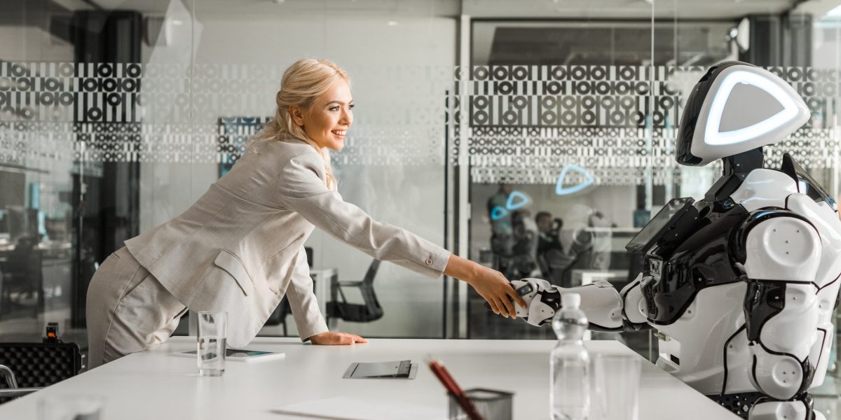 smiling businesswoman shaking hands with robot sitting at desk in meeting room